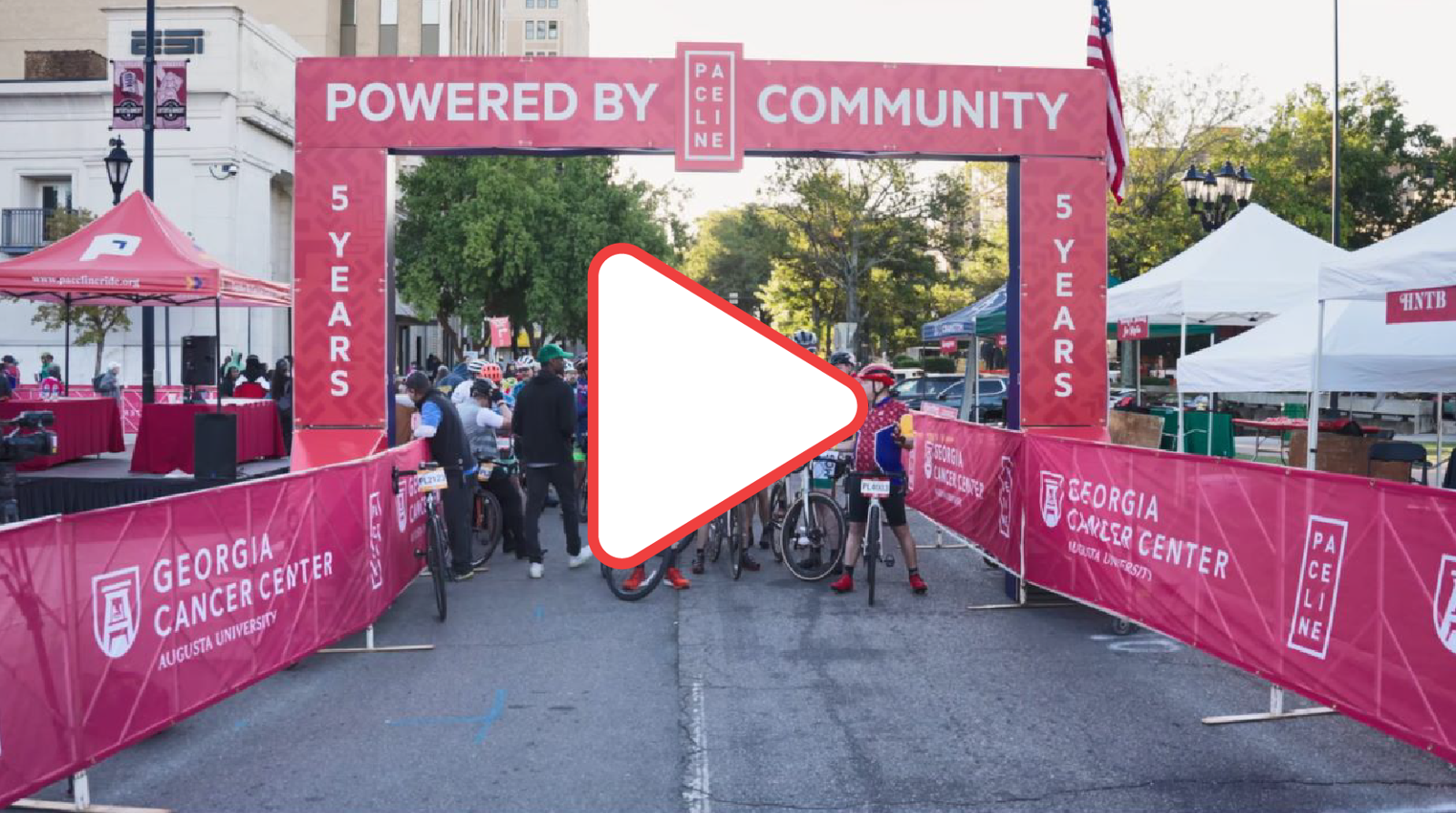 A group of cyclists lining up under a start truss with the words Powered By Community. There is also a play button overtop indicating a link.