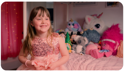 Miracle Child Harper smiles as she tells her accident story from her bed, surrounded by stuffed animals, wearing a sequined pink dress